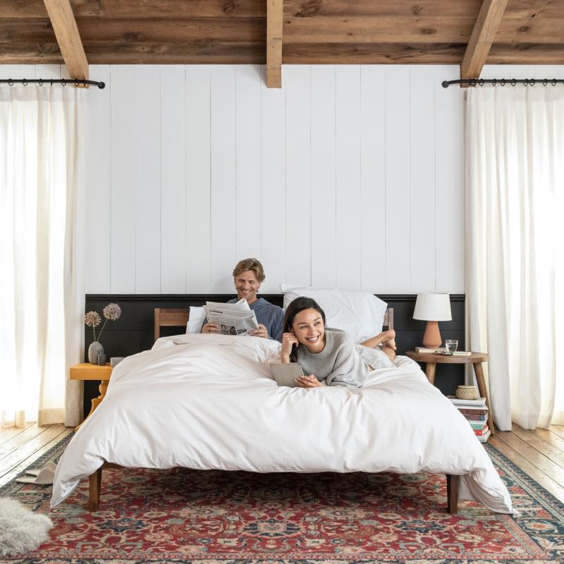 A new-style lifestyle image in a warm, lived-in space. A couple lies on the bed reading and laughing.